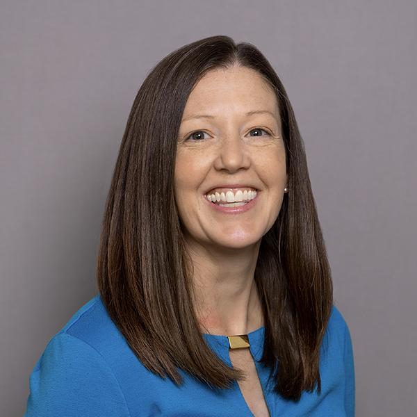 Professional portrait of a smiling woman, wearing a blue shirt against a grey background.